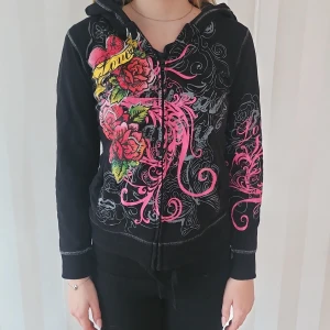 Svart hoodie med blommigt mönster - Säljer en cool svart hoodie med färgglatt blommigt mönster i rosa och gult. Den har en dragkedja framtill och luva. Perfekt för en avslappnad stil med lite edge. Är i barnstorlek men motsvarar en 34.