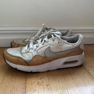 Nike sneakers i beige och vit - Säljer ett par Nike sneakers i beige och vit med snörning. De har en klassisk design med en synlig luftkudde i sulan för extra komfort. Skorna är i bra skick men har några tecken på användning. Perfekta för vardagsbruk eller träning!