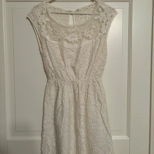 Vit spetsklänning - Säljer en vit spetsklänning med många detaljer framtill i bröstet. Den är draperad i midjan vilket gör att figuren framhävs fint. Ryggen har guldknappar. Den slutar i lårlängd. Storlek XXS.