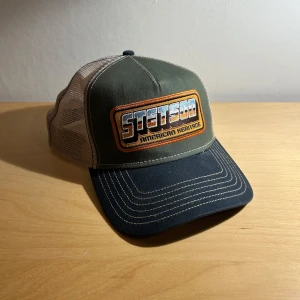 Grön och beige Stetson keps   - Snygg keps från Stetson i grönt och beige med ett stort broderat märke framtill. Kepsen har en klassisk truckerdesign med mesh på baksidan för extra ventilation. Perfekt för en avslappnad stil. Skicket ör grymt utan några defekter!