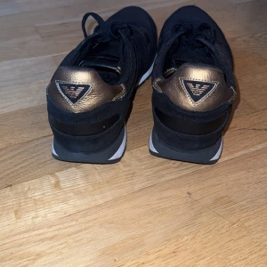 Svarta sneakers med guldlogga från Emporio Armani - Snygga svarta sneakers från Emporio Armani med en iögonfallande guldlogga på sidan och hälen. Skorna har snörning och en vit sula som kontrasterar snyggt mot det svarta. Perfekta för en stilren look.