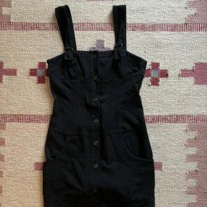 Svart klänning med knappar - Snygg svart klänning med knappar framtill och justerbara axelband. Perfekt för en stilren look. Klänningen har en figurnära passform och praktiska fickor.