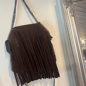 Brun axelväska med fransar kedjeväska - Snygg brun axelväska med coola fransar och kedjedetaljer. Perfekt för att ge din outfit en bohemisk touch. Väskan har en unik design med en kedja som axelrem. Har aldrig använt väskan
