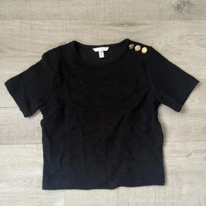Svart kortärmad topp med guldknappar - Svart topp från H&M med korta ärmar och  guldknappar på axeln. Strl S. Aldrig använd 