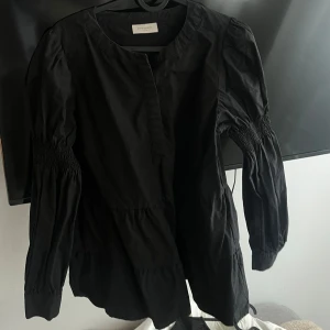 Svart blus  från H&M - Snygg svart blus från H&M med långa ärmar och smockdetaljer vid ärmarna. Perfekt för en stilren look. Blusen  har en rund halsringning och en avslappnad passform.