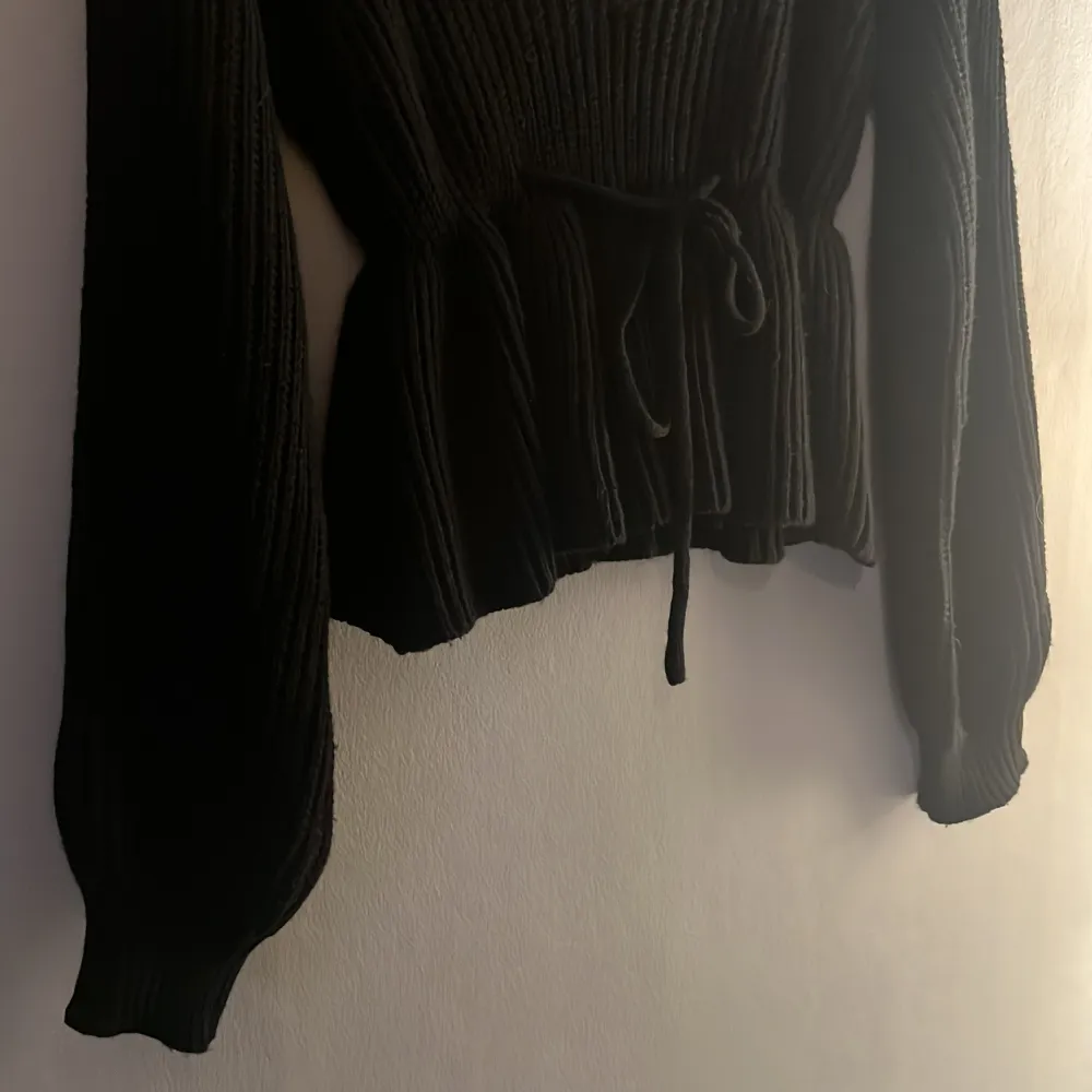 Snygg svart ribbad tröja med långa ärmar och knyt i midjan. Den har en v-ringad design med fina detaljer som ger en stilren look. Perfekt för att hålla sig varm och trendig.. Neuletakit & Villapaidat.