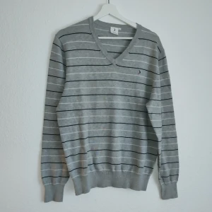 Grå randig tröja från Boomerang - Säljer en stilren grå tröja från Boomerang med svarta ränder och en liten broderad logga på bröstet. Tröjan har en v-ringad hals och långa ärmar, perfekt för en avslappnad look.