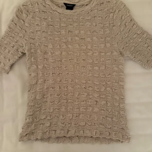 Beige topp från Lindex - Säljer en beige topp från Lindex med ett unikt texturerat mönster. Toppen har korta ärmar och en rund halsringning. Perfekt för att ge din outfit en intressant touch. Passar bra till både jeans och kjol🤍