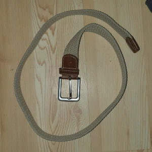 Beige flätat bälte med läderdetaljer - Snyggt beige flätat bälte med bruna läderdetaljer och en silverfärgad spänne. Perfekt för att ge en stilren touch till din outfit. Bältet är flexibelt och passar de flesta storlekar.