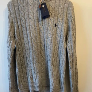 Grå stickad tröja från Ralph Lauren halv zip - Säljer en stilren grå stickad tröja från Ralph Lauren med klassiskt flätmönster. Tröjan har en diskret broderad logga på bröstet och en dragkedja vid halsen. Perfekt för kyligare dagar!
