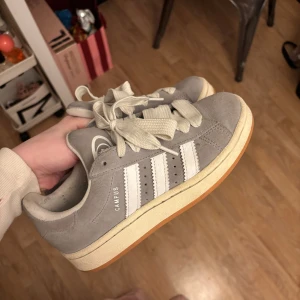 Grå Adidas Campus sneakers - Säljer ett par grå Adidas Campus sneakers i mocka med vita detaljer och snörning. Dem är inte använda så mycket utan skulle gissa på använda 10 ggr, skicket är helt okej, inte helt nya men inte förstörda. Har inte testat att rengöra dem själv så de kan man ju göra för att få till dem ännu mer ☺️