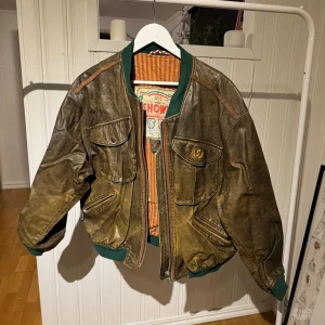 Brun skinnjacka från Showa - Säljer en cool brun skinnjacka från Showa med gröna detaljer. Jackan har en vintage look med dragkedja och stora fickor framtill. Insidan har ett randigt mönster i orange och vitt. Perfekt för dig som vill ha en unik stil!