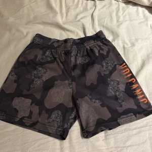 Camouflage shorts med tryck - Snygga shorts i camouflage-mönster med orange tryck på sidan. Perfekta för en avslappnad stil. Elastisk midja för extra komfort.