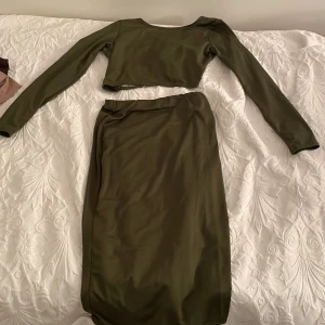 Olivgrönt set med långärmad topp och kjol - Snyggt olivgrönt set med en långärmad croppad topp och matchande kjol. Toppen har en omlott-design med knytning i midjan, vilket ger en trendig look. Perfekt för att skapa en stilren outfit.
