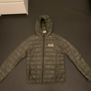 Grön pufferjacka från EA7 - Snygg grön pufferjacka från EA7 med dragkedja och huva. Perfekt för kyligare dagar. Jackan har en quiltad design och är långärmad. EA7-loggan syns på bröstet. Jackan funkar alla årstider. 