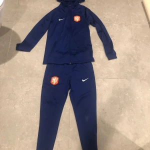 Blå träningsjacka från Nike - Snygg blå träningsjacka från Nike med huva och dragkedja. Jackan har ett Nederländsk emblem på bröstet och en vit Nike-logga. Perfekt för sportiga aktiviteter och att hålla sig varm.