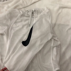 Vit meshtröja från Nike - Säljer en vit meshtröja från Nike med dragkedja och ett stort svart Swoosh-märke på framsidan. Perfekt för sportiga tillfällen eller som ett stilrent lager. Tröjan har långa ärmar och är tillverkad i ett lätt och luftigt material.