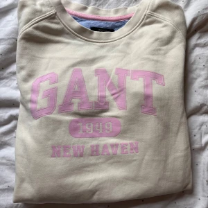Beige sweatshirt från GANT - Säljer en beige sweatshirt från GANT med rosa text 'GANT 1949 NEW HAVEN' på framsidan. Tröjan har en rund halsringning och är perfekt för en avslappnad stil. Passar barn i storlek 146/152.