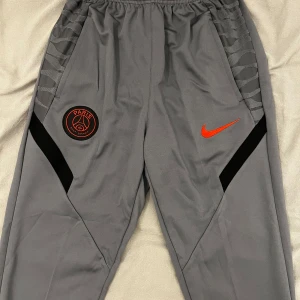 Grå träningsbyxor från Nike - Snygga grå träningsbyxor från Nike med Paris Saint-Germain-logga. Byxorna har en elastisk midja och svarta detaljer längs sidorna. Perfekta för sport och avslappnade dagar.  Koftan finns även om intresserad 