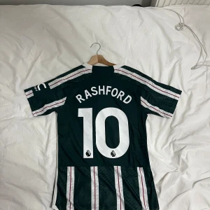 Grön och vit randig fotbollströja, manchester united  - Säljer en grön och vit randig fotbollströja från Adidas med spelarnamnet 'Rashford' och nummer 10 på ryggen. Tröjan har korta ärmar och klubbmärket på bröstet. Perfekt för fotbollsfans!helt ny 