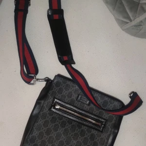 Svart axelväska från Gucci - Snygg svart axelväska från Gucci med det klassiska monogrammönstret. Väskan har en justerbar axelrem i rött och blått samt en praktisk dragkedja framtill. Perfekt för att bära dina nödvändigheter med stil.