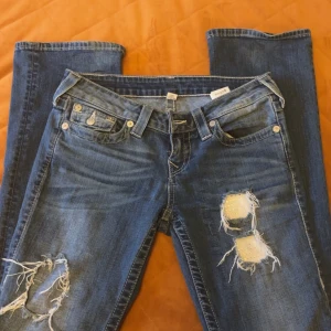 Blå jeans med slitningar - Snygga blå jeans från True Religion med slitningar på framsidan. Klassisk femficksdesign och knappgylf. Perfekta för en avslappnad stil med en touch av edge.