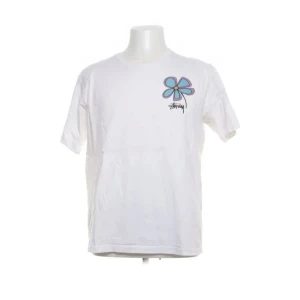 Vit t-shirt med blomtryck från Stüssy - Säljer en vit t-shirt från Stüssy med ett stort färgglatt blomtryck på både fram- och baksidan. T-shirten har korta ärmar och är tillverkad i bomull. Perfekt för en avslappnad stil.