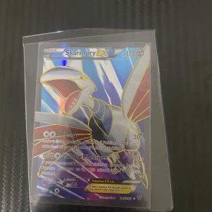 Skarmory EX Pokémonkort - Säljer ett Skarmory EX Pokémonkort med 170 HP. Kortet har attacker som Joust och Tailspin Piledriver. Det är en holografisk design med en bild av Skarmory i bakgrunden.