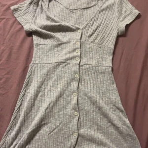 Grå ribbad klänning från Pull&Bear - Säljer en fin och söt grå ribbad klänning från Pull&Bear. Klänningen har korta ärmar och knappar framtill som ger en snygg detalj. Perfekt för en avslappnad dag på sommaren.