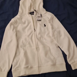 Vit hoodie från Ralph Lauren - Säljer en stilren vit hoodie från Ralph Lauren med dragkedja och broderad logga på bröstet. Den har en justerbar huva och praktiska fickor framtill. Perfekt för en avslappnad look. 600 kr helt ny 