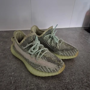 Adidas Yeezy sneakers - Snygga Adidas Yeezy sneakers i en mix av beige och svart med en unik stickad design. Skorna har en ribbad sula i ljusgrön färg och snörning i matchande ton. Perfekta för dig som vill ha en stilren och modern look.