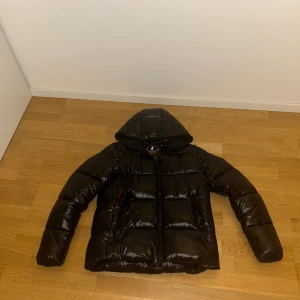 Svart pufferjacka från Save The Duck - Snygg svart pufferjacka från Save The Duck med glansig finish. Jackan har en huva och är märkt med '100% Animal Free'. Perfekt för kyliga dagar med stil. Endast använd 3 gånger och är så gott som ny!