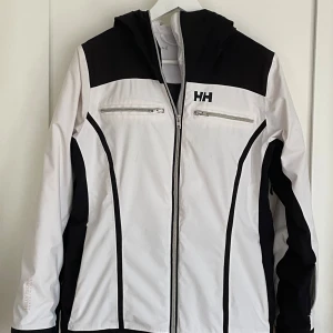 Vit och svart skidjacka dam i strl M från Helly Hansen - Snygg vit och svart damskidjacka från Helly Hansen med dragkedja och huva i strl M. Jackan har två dragkedjefickor framtill och ett stilrent märke på bröstet. Jackan är vind-och vattentät.