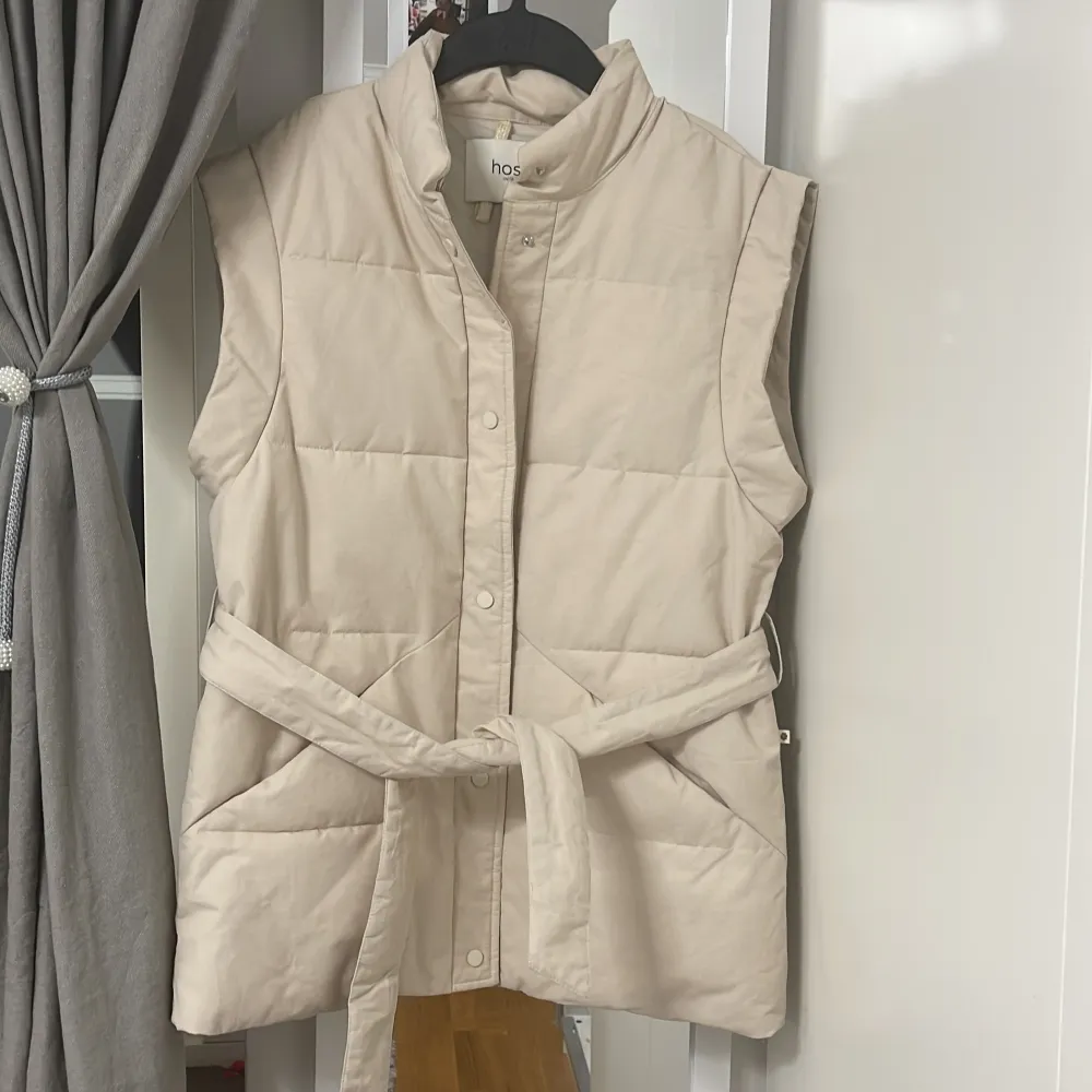 Säljer en stilren beige quiltad väst från Hoss Intropia. Västen har en hög krage och knäpps med knappar framtill. Den har ett bälte i midjan för en snygg passform. Perfekt för lager-på-lager under kyligare dagar.. Takit.