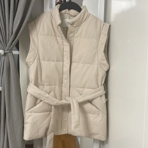 Beige quiltad väst från Hoss Intropia - Säljer en stilren beige quiltad väst från Hoss Intropia. Västen har en hög krage och knäpps med knappar framtill. Den har ett bälte i midjan för en snygg passform. Perfekt för lager-på-lager under kyligare dagar.