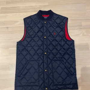 Mörkblå quiltad väst från Ralph Lauren -  Funkar både för män som har S och M! Snygg mörkblå quiltad väst från Polo Ralph Lauren med röd insida och broderad logga på bröstet. Västen har knappar framtill och är perfekt för en stilren look. Passar bra till både casual och mer uppklädda tillfällen.