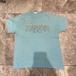 Blå t-shirt från Gildan - Säljer en blå t-shirt från Gildan med texten 'Marathon Florida Est. 1821' tryckt på framsidan. T-shirten är kortärmad och tillverkad i bomull, perfekt för en avslappnad stil.