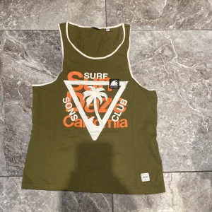 Olivgrön linnetopp från Quiksilver - Säljer en olivgrön linnetopp från Quiksilver med tryck i vitt och orange. Toppen har ett surf-inspirerat motiv med text och palmträd. Perfekt för sommarens äventyr!