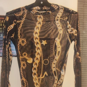 Svart mesh body med kedjemönster från Boohoo - Säljer en svart mesh body från Boohoo med ett iögonfallande kedjemönster i guld. Toppen har långärmade ärmar och en tight passform, perfekt för en trendig look.