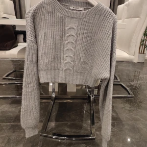 Grå stickad tröja från NA-KD - Säljer en snygg grå stickad tröja från NA-KD. Tröjan är croppad med ett flätat mönster framtill och har långa ärmar. Perfekt för en stilren look.