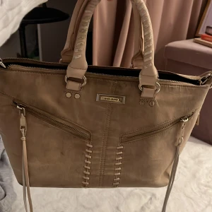 Beige handväska från river Island  - Snygg beige handväska från river Island med dragkedja och coola detaljer. Perfekt för både vardag och fest! Den har två handtag och en stilren design som passar till allt. Väskan är i syntetmaterial och har en praktisk storlek för alla dina nödvändigheter.