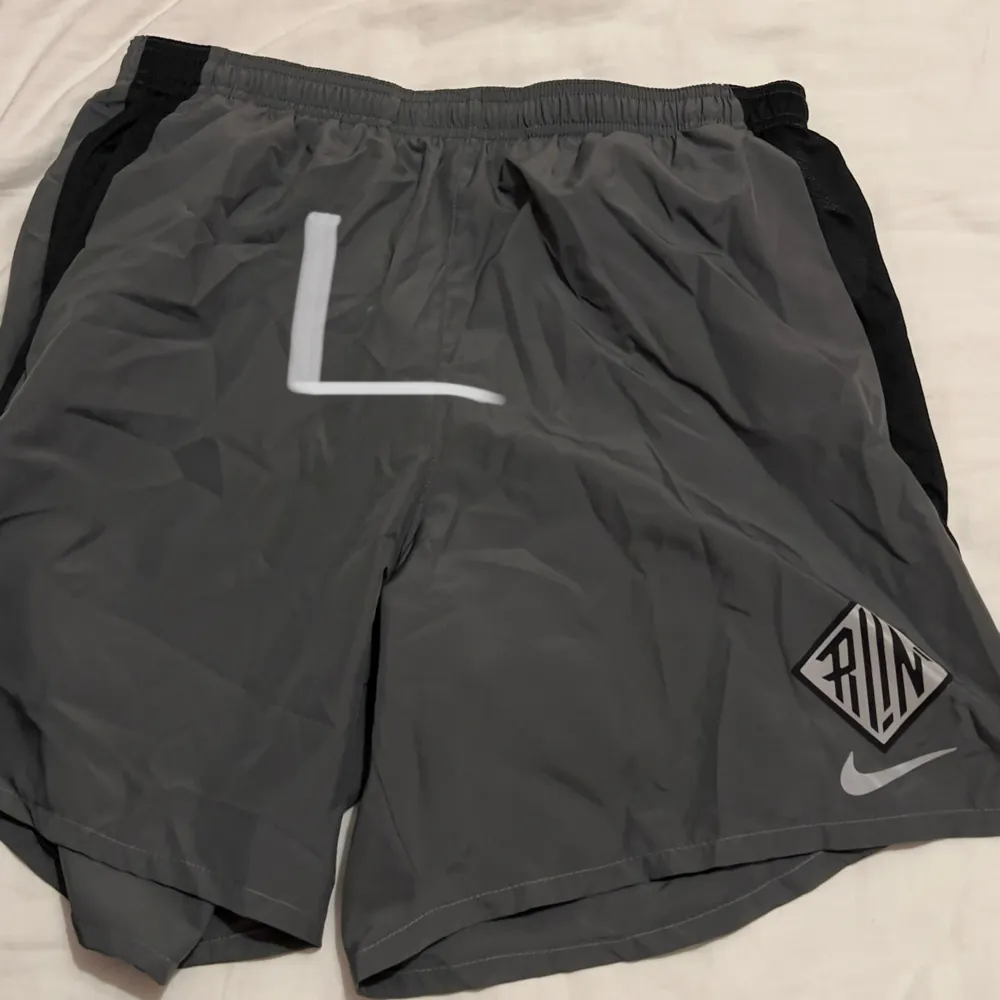 Säljer ett par grå träningsshorts från Nike med svart detalj på sidorna och en vit logga. Perfekta för löpning eller gymmet. De har en elastisk midja för bättre passform och är gjorda i ett lätt och bekvämt material.. Shorts.