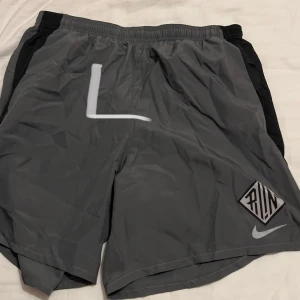 Grå träningsshorts från Nike - Säljer ett par grå träningsshorts från Nike med svart detalj på sidorna och en vit logga. Perfekta för löpning eller gymmet. De har en elastisk midja för bättre passform och är gjorda i ett lätt och bekvämt material.