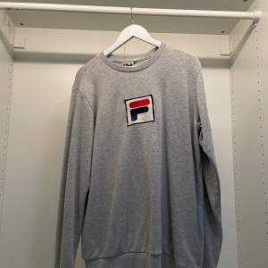 Säljer en stilren grå sweatshirt från Fila i storlek L. Tröjan har en klassisk rund hals och ett stort Fila-logotyp på bröstet. Perfekt för en avslappnad stil eller träning. Den är i bra skick och superbekväm att ha på sig. Passar både till vardags och sportiga aktiviteter.