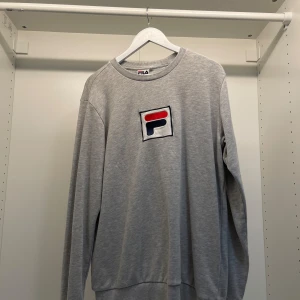 Grå sweatshirt från Fila - Säljer en stilren grå sweatshirt från Fila i storlek L. Tröjan har en klassisk rund hals och ett stort Fila-logotyp på bröstet. Perfekt för en avslappnad stil eller träning. Den är i bra skick och superbekväm att ha på sig. Passar både till vardags och sportiga aktiviteter.