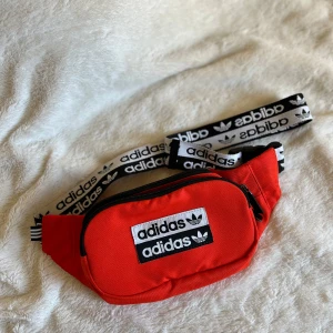 Röd magväska från Adidas - Säljer en snygg röd magväska från Adidas med justerbart band och dragkedja. Perfekt för att hålla dina saker säkra och nära till hands. Orange/red colour