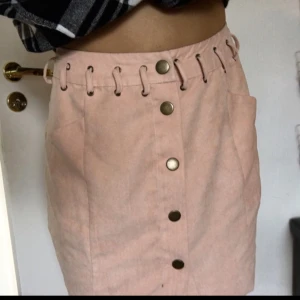  - Ljusrosa kjol i fejkmocka! Brons/guldig knäppning fram och knytdetaljer vid midjan fram. Den är uppsydd och något insydd men fortfarande stl 36 och för stor för mig. Omsyningen går att sprätta. Kan fraktas då köparen står för kostnaden!