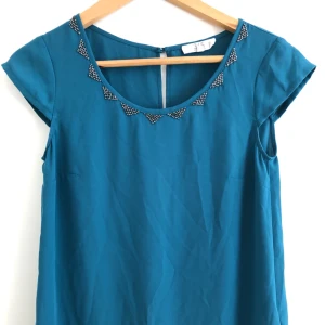  - Tunn topp i petrol/turkos med kort ärm och pärlor längs urringningen. Vidare nedtill. Snygg!