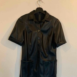  - Helt oanvänd svart skinnklänning med knappar och två fickor framtill. 