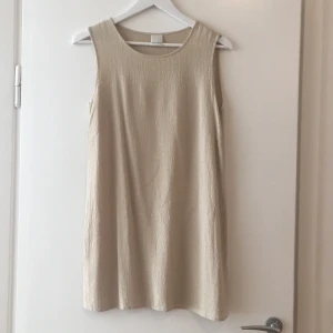 Beige klänning, 90-tal - En beige klänning från 90-talet. Materialet är polyester med beiga broderier så det ser lite ut som ormskinn. Den är ganska kort o går lite ovanför mitten på låret. Finns två slitsar på höger o vänster sida längst ner.  
